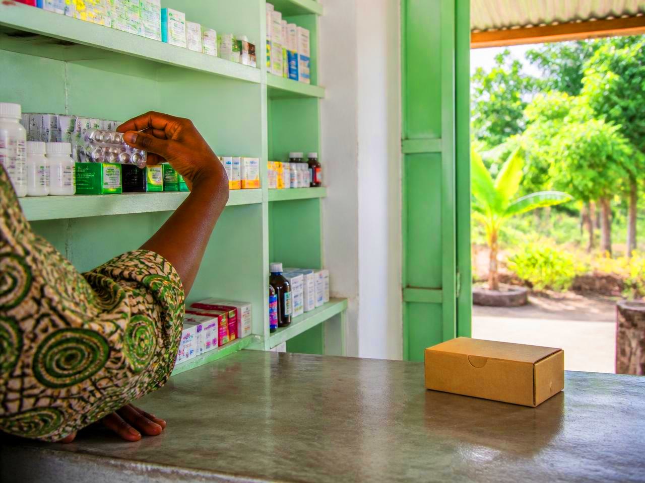 A hand reaching to a shop shelf for a blister strip of tablets, an unopened rapid malaria test box on the counter to one side