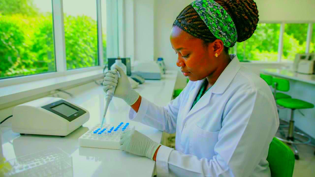 A laboratory technician holding a pipette over a rack of labelled sample vials, an open notebook beside the rack