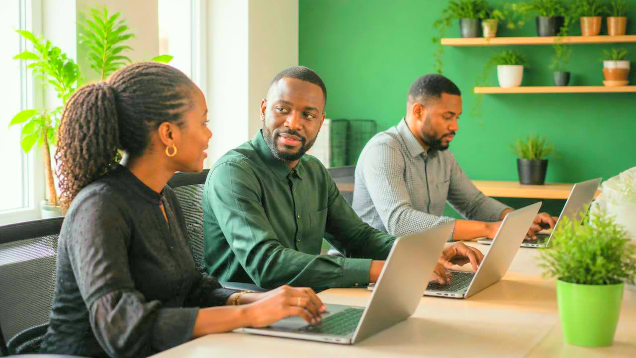 Two programme officers in an office comparing a printed stock report against a laptop, ring-bound files and a desk fan beside them